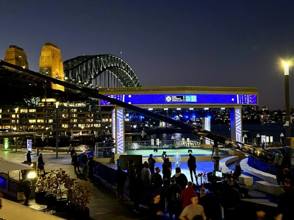 Night scene captures a stage setup for FIFA Women's World Cup in Sydney, with onlookers and iconic architecture in the background.