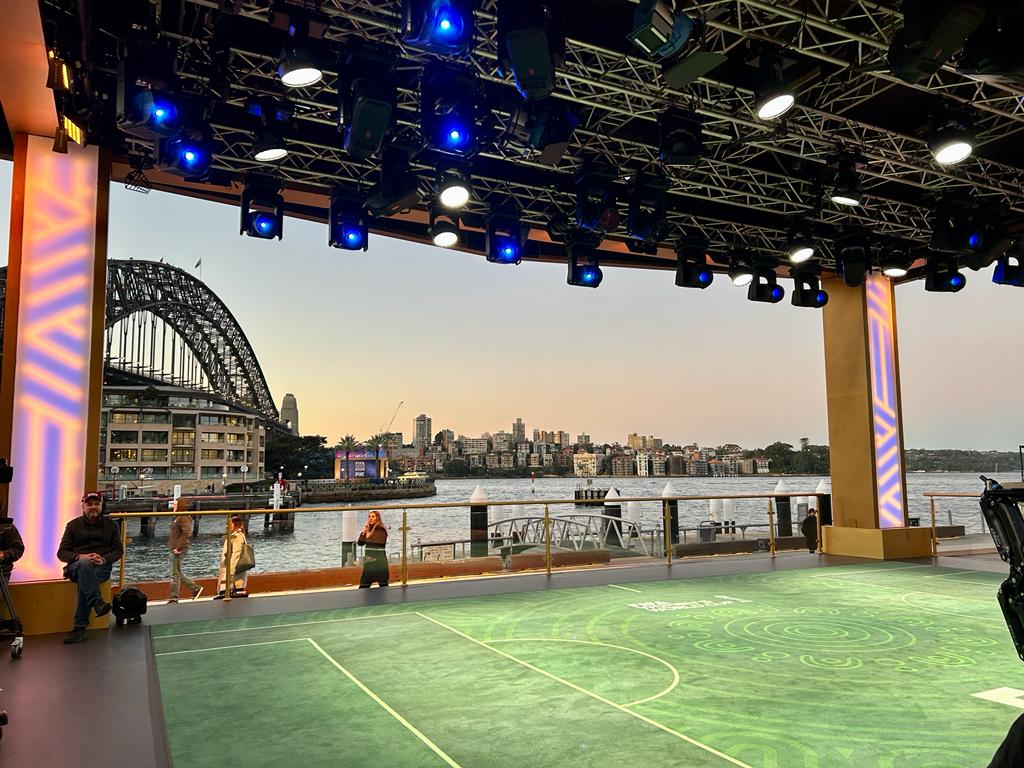 Outdoor shot of people on a patio overlooking the water and the Sydney Harbour Bridge, with a stage set and lighting rig visible.