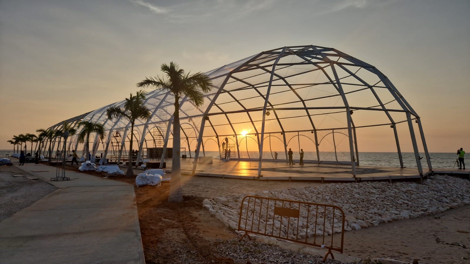 Large clear event tent structure on a beach at sunset, with silhouettes of people working inside and palm trees along the side.