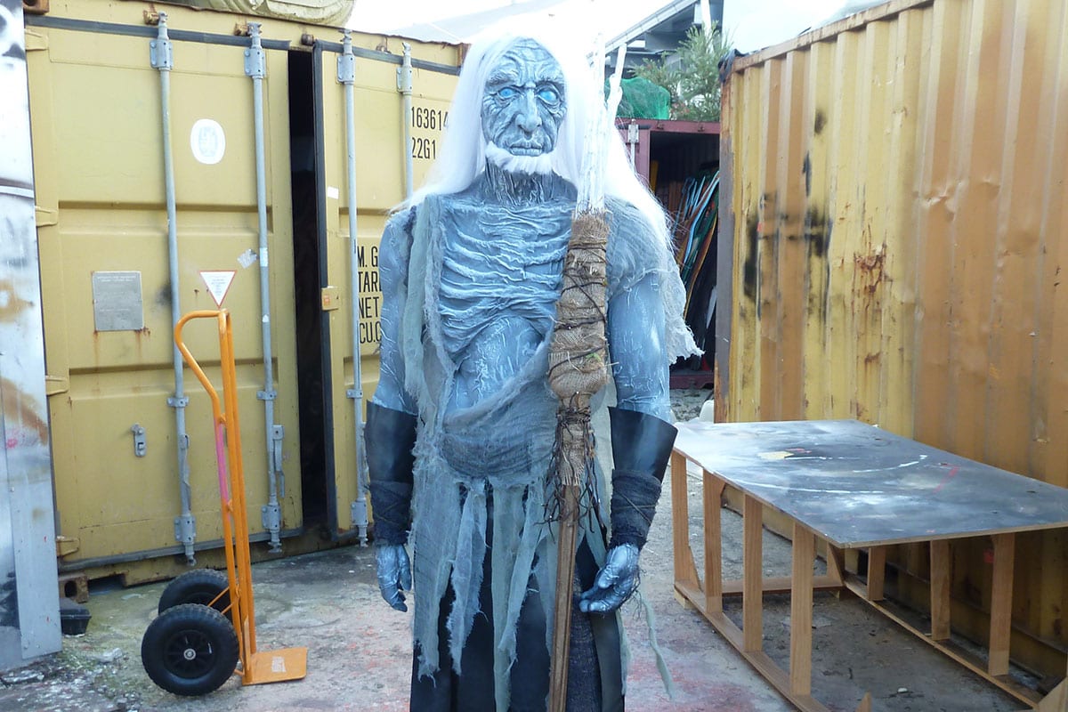 Costumed person with white hair and pale blue skin holds a staff, standing outdoors near a yellow shipping container on a cloudy day.