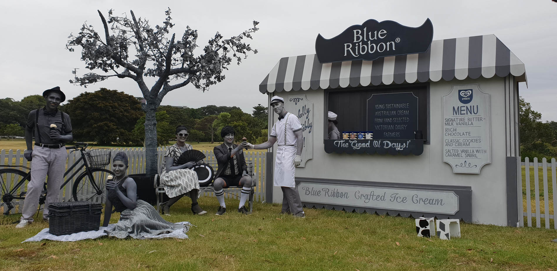 Retro-style picnic scene with actors in gray-scale makeup, featuring an ice cream stand and vintage props in an outdoor setting.