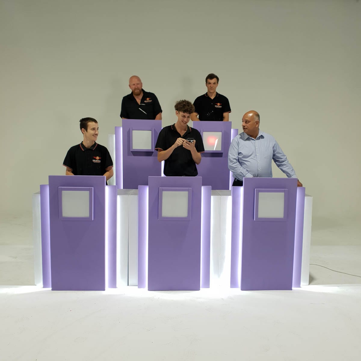 Five people stand behind individual podiums in a studio setting. One person is looking at a phone.