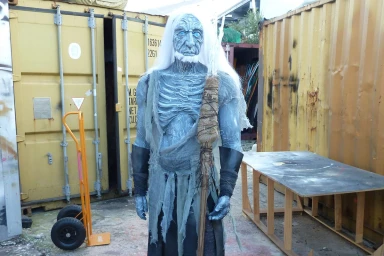Full-body costume of a blue-skinned fantastical creature with long white hair holding a staff, set outdoors near industrial storage containers.