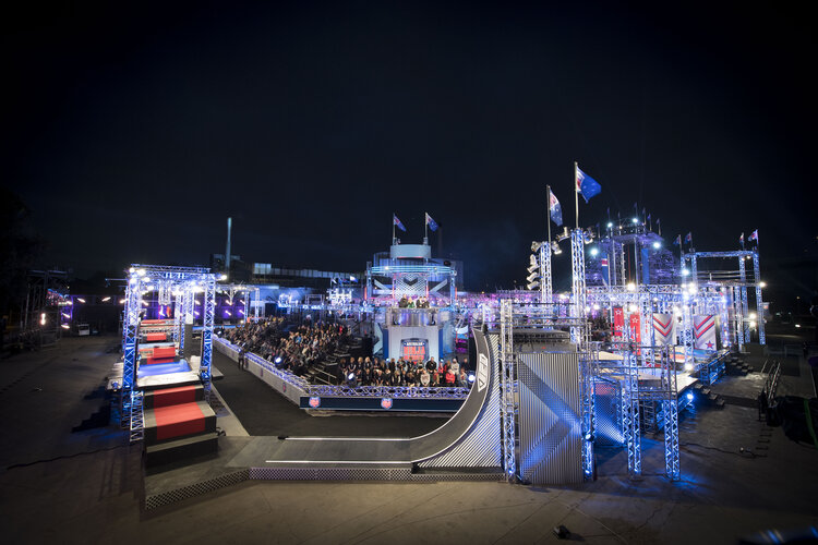 Outdoor nighttime shot of an elaborate obstacle course structure with a large crowd of people in the background under blue lighting.
