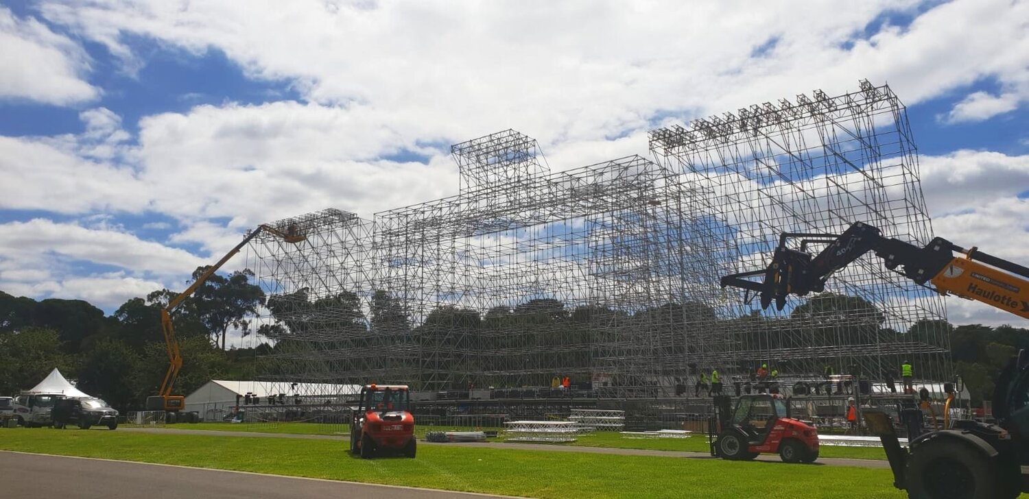 Outdoor daytime shot featuring a large metal scaffolding structure under a cloudy blue sky, with heavy machinery on the grassy ground.