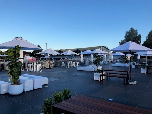 Outdoor event space featuring white umbrellas, cube seating, and high-top tables under a clear sky in a modern, open-air setup.