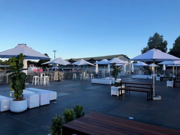 Outdoor event space featuring white umbrellas, cube seating, and high-top tables under a clear sky in a modern, open-air setup.