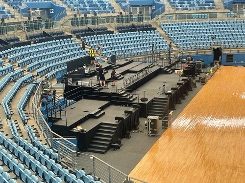 Event setup in arena features black stage platform, lighting rigs, audio equipment for upcoming live show, and rows of empty blue seating.