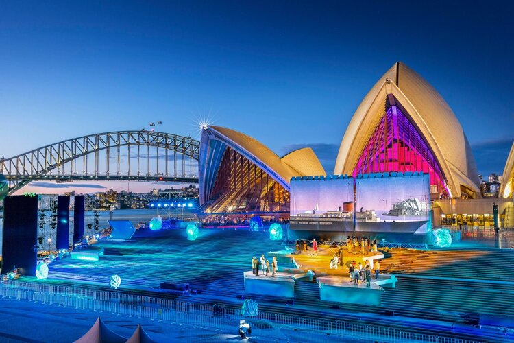 Sydney Opera House illuminated at night during a festival, with Sydney Harbour Bridge in the background and a crowd of people watching.