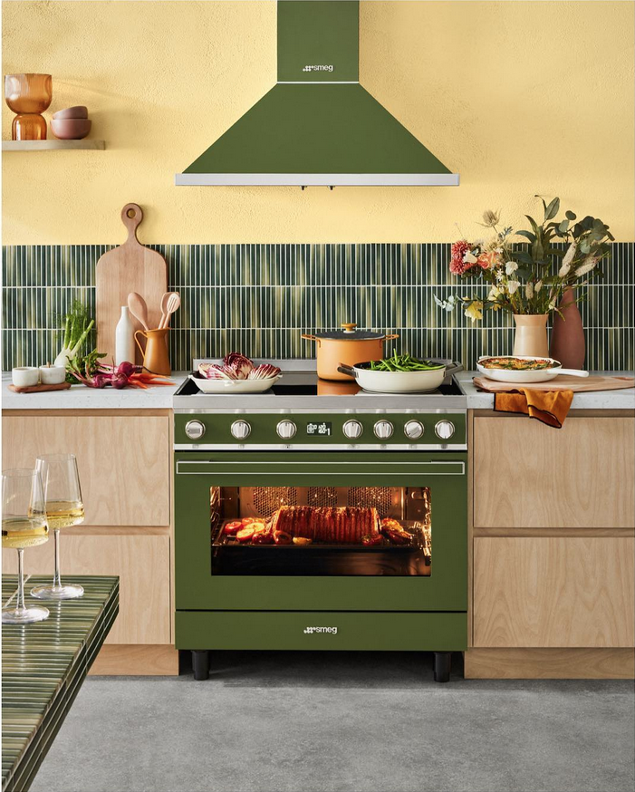 Kitchen interior with a green Smeg oven, range hood, assorted cookware, dishes, produce, and glasses of white wine on counter and table.