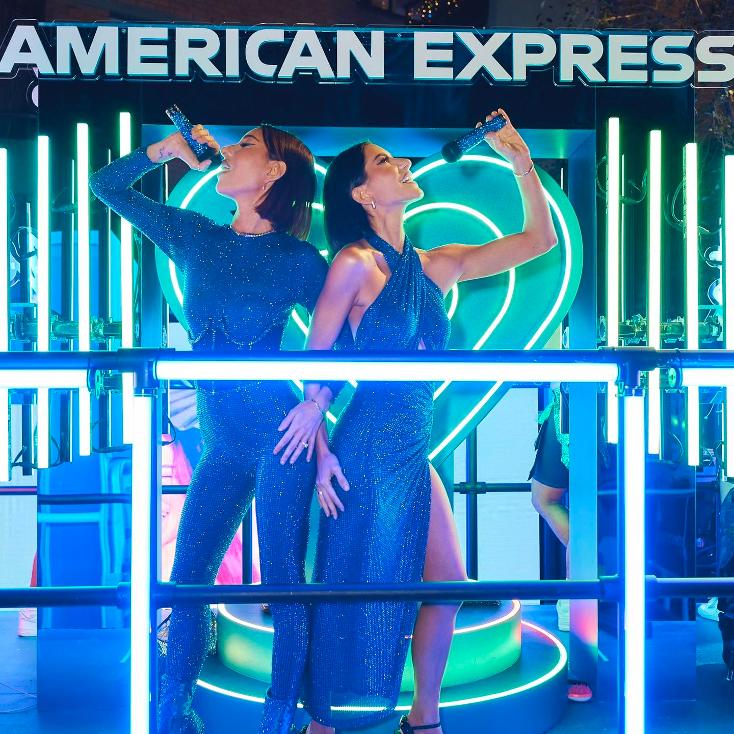 Two women singing into microphones on a stage with blue lighting and an American Express sign in the background. Evening shot.