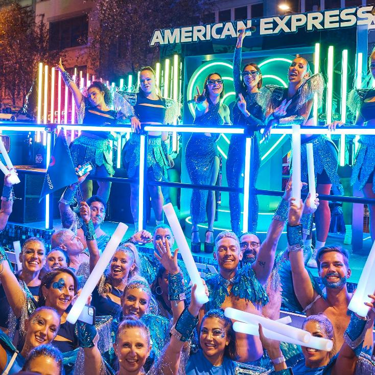Large group of people celebrate on a float with blue lights and American Express signage, creating a vibrant and festive atmosphere.