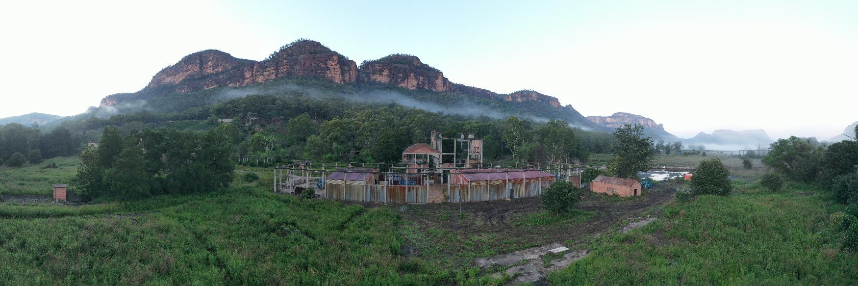Panorama of an abandoned building amidst lush greenery, mountains in the distance, under a soft, overcast sky. Serene yet haunting landscape.