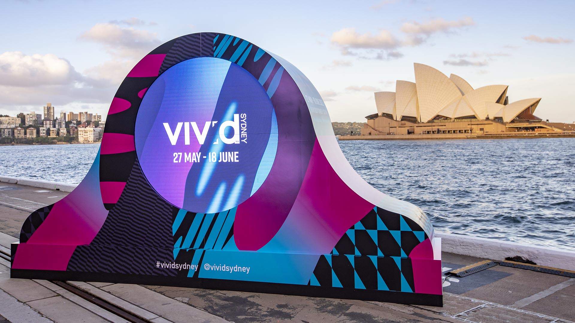 Vivid Sydney promotional installation on a pier with the Sydney Opera House visible in the background under a blue sky.