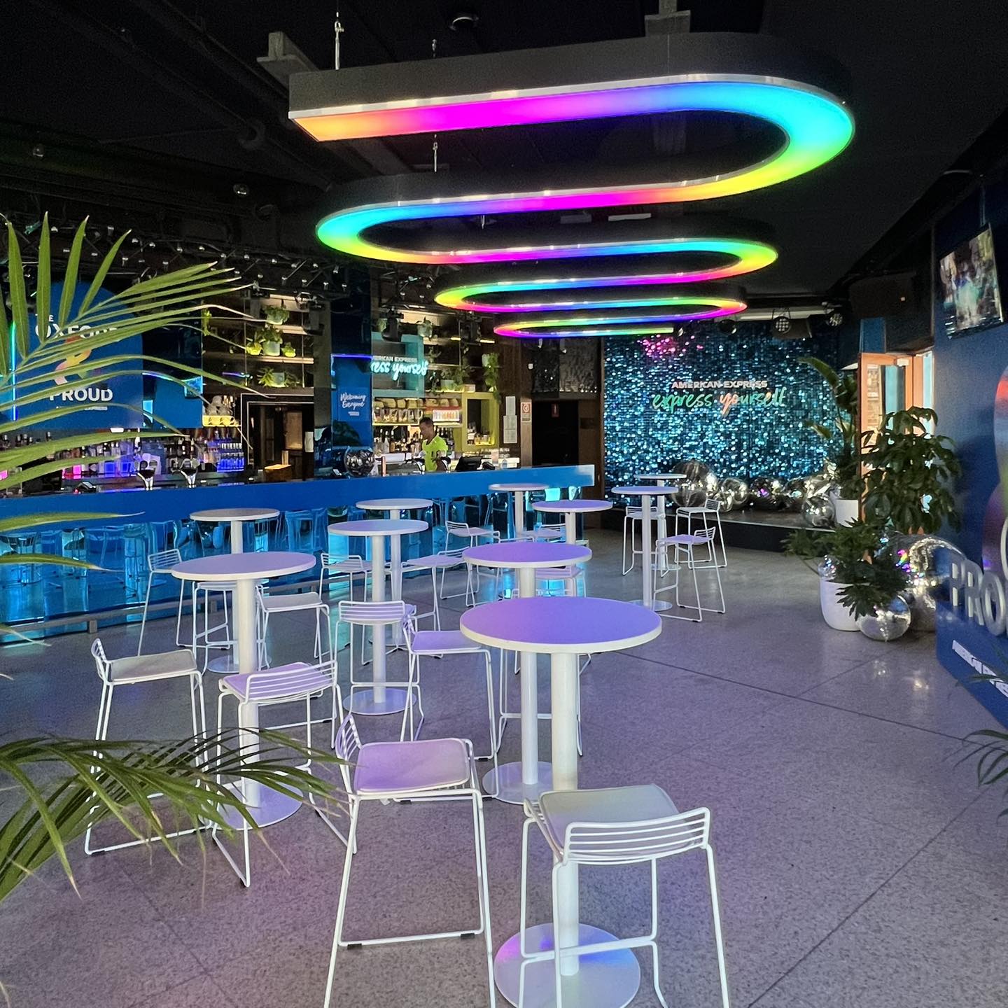 Interior view of a bar with colorful LED lighting, white tables and chairs, lush potted plants, and a lively, modern ambiance.