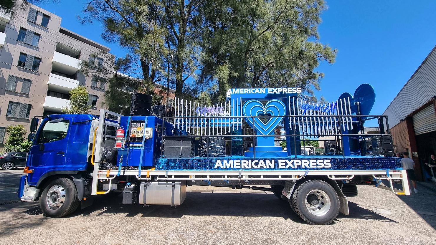 Blue custom truck with American Express branding and a heart-shaped decoration, parked outdoors under a bright sky, with buildings in the background.