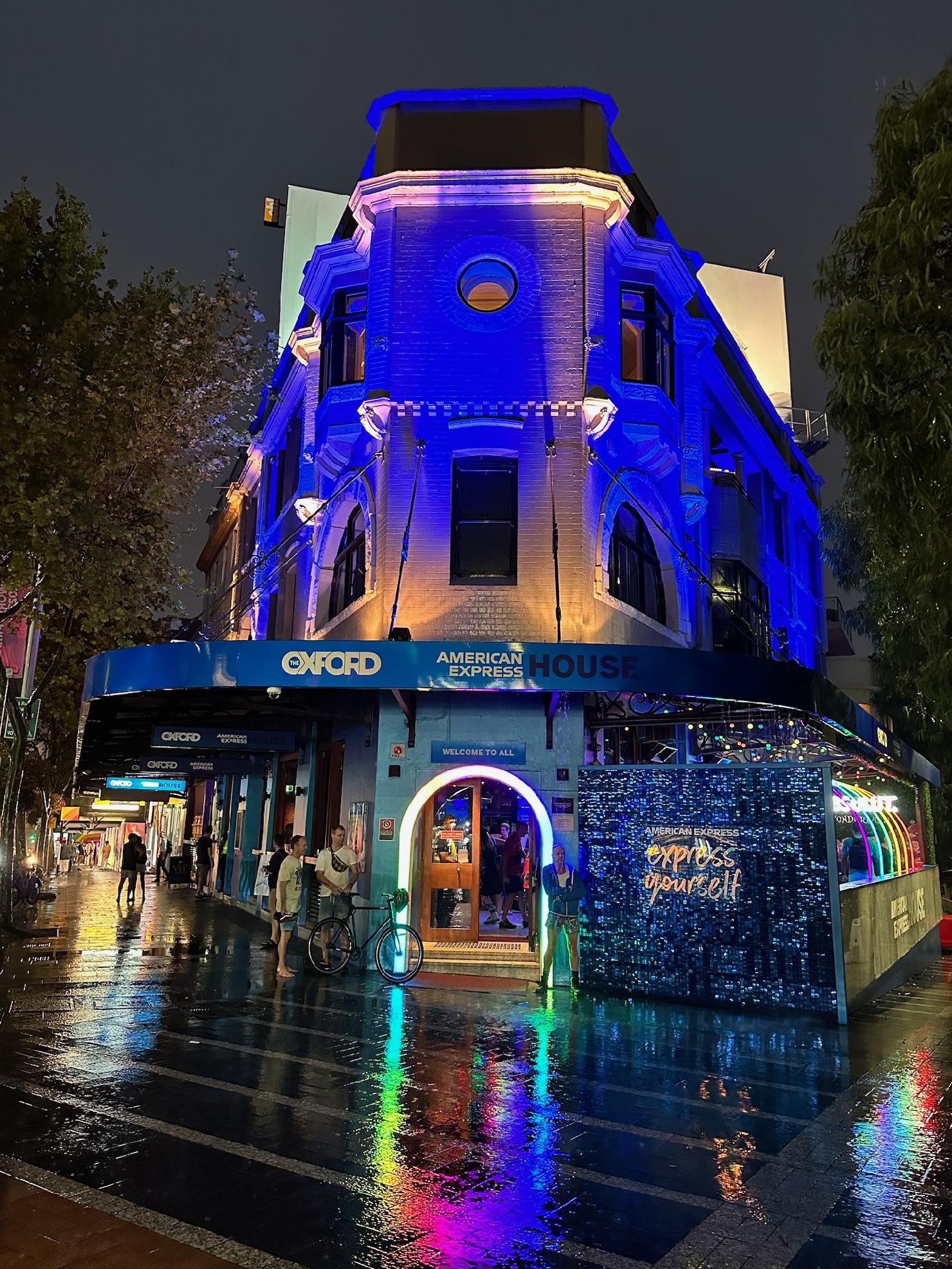Illuminated American Express House at night. Wet pavement reflects colorful lights. Two pedestrians, a bicycle, and building details visible.