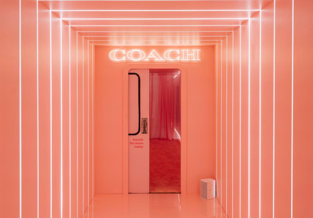 A photo booth is framed by LED lights, creating a tunnel effect in a peach-colored room, highlighting the Coach brand name at the entrance.