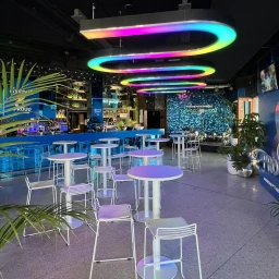 Modern bar interior featuring white high-top tables, wire-frame stools, and glowing rainbow LED ceiling lights by Wildsets.