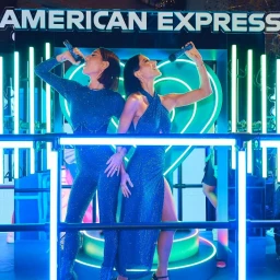 Two women in sparkling blue outfits singing into microphones on an illuminated stage with neon lights and American Express branding.