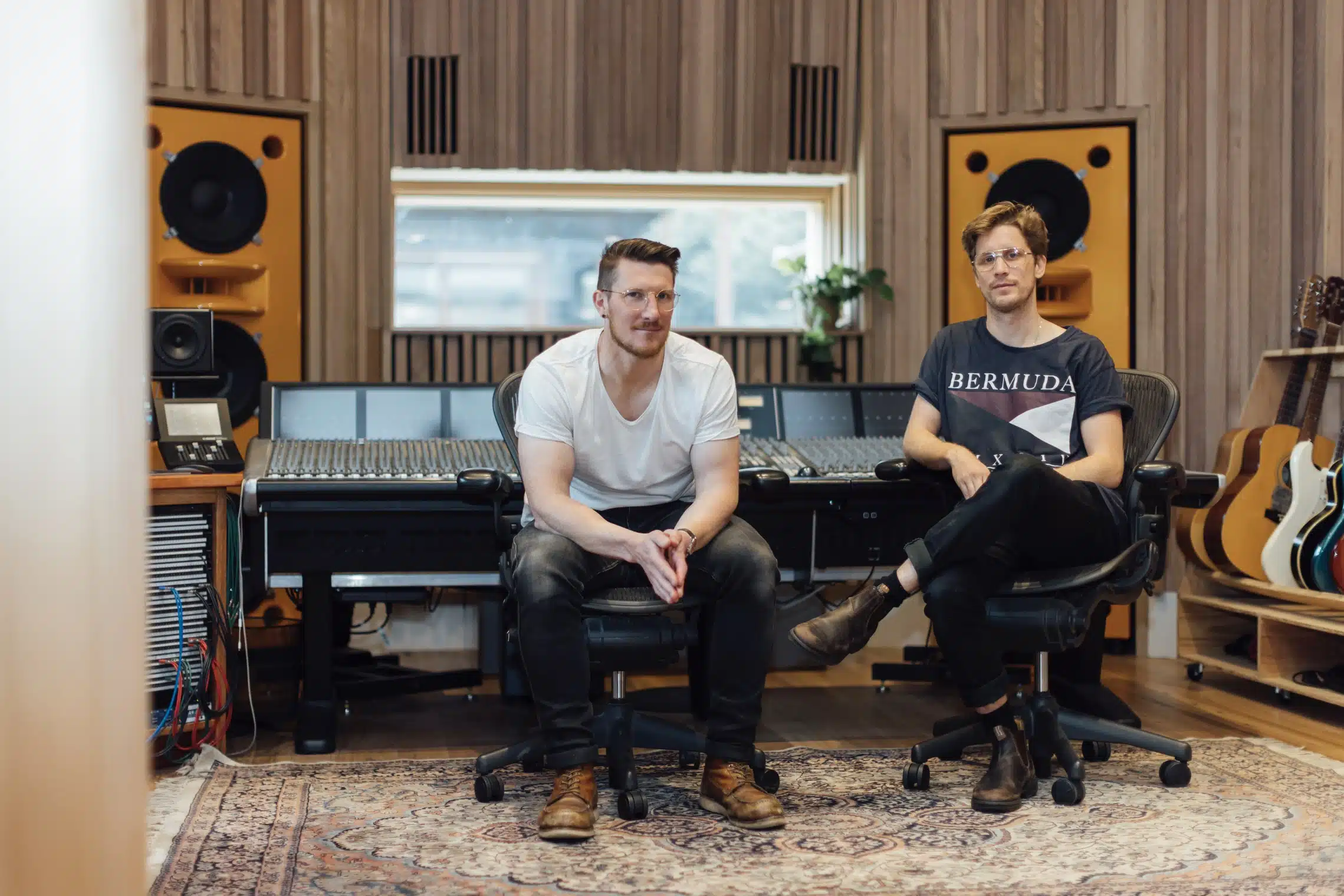 Two adult men are sitting in a music studio with a large audio mixing board, speakers, microphones, guitars, and soundproofing visible.