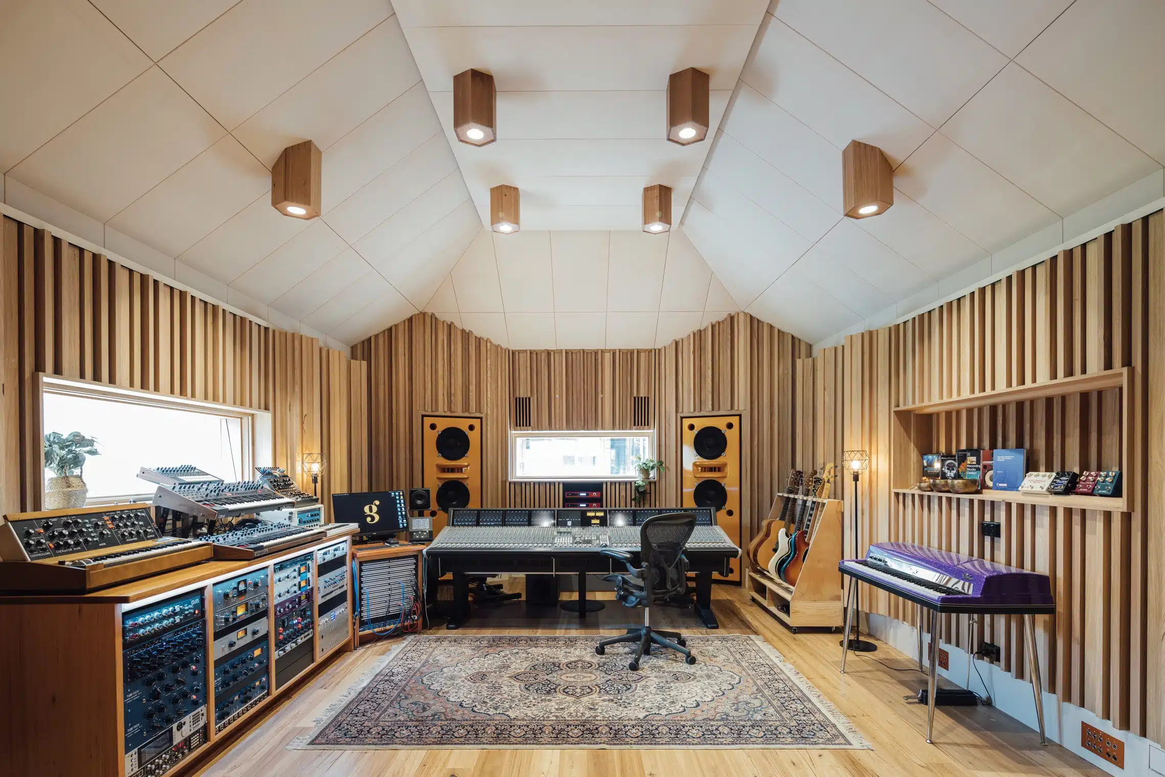 A well-equipped music studio with wood-paneled walls, featuring a mixing console, synthesizers, speakers, and guitars in a professional setting.