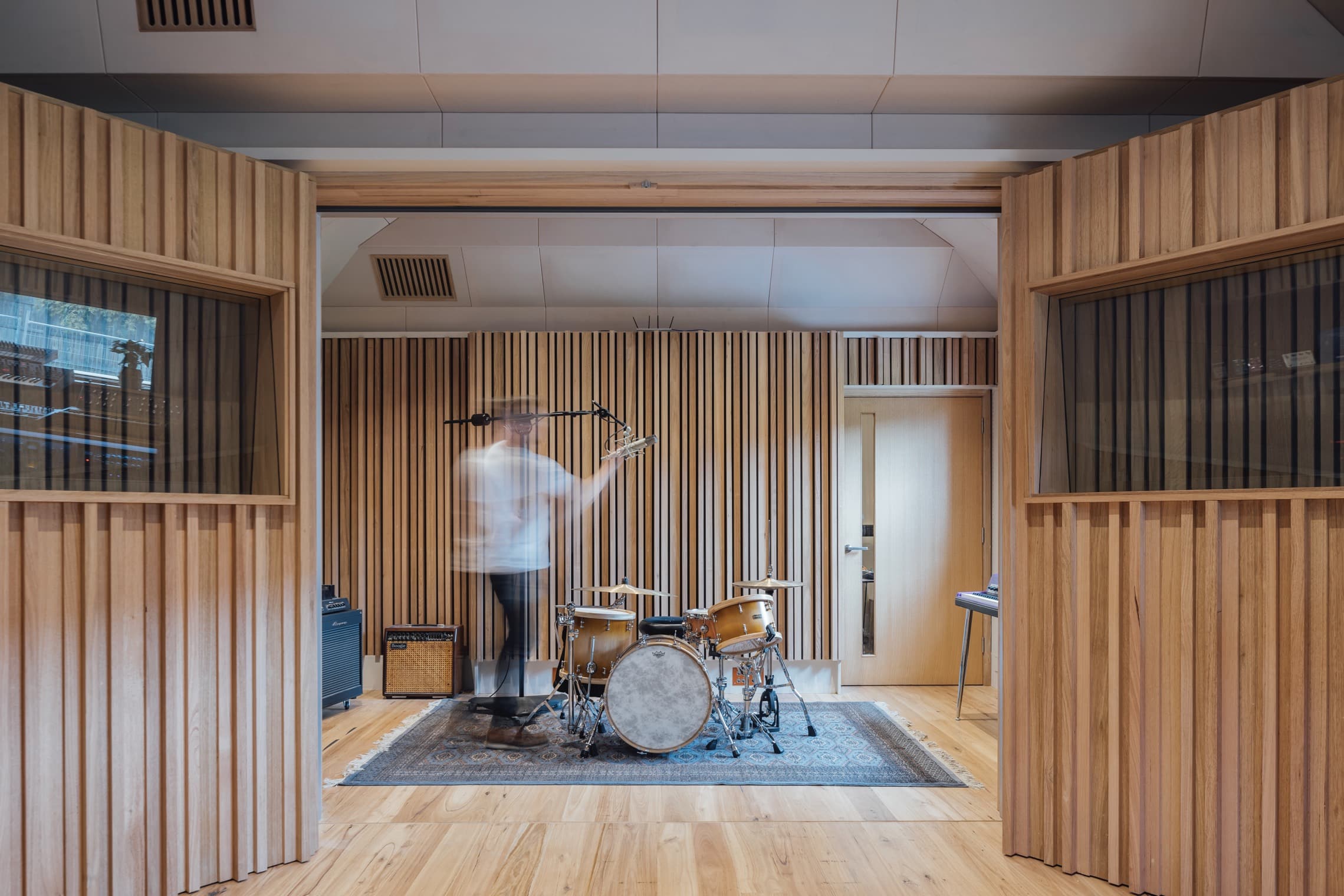 Man playing drums in a wood-paneled music recording studio. The room has a drum set, amplifier, microphone, and a blue patterned rug.