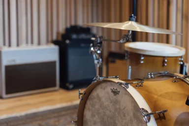 Close-up of a wooden drum kit in a recording studio with acoustic wall panels and guitar amplifier