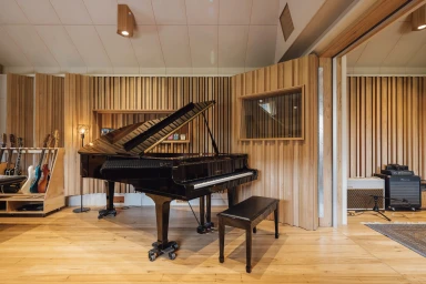A black grand piano and matching bench inside a modern Melbourne audio production studio with wooden slatted walls and acoustic paneling.