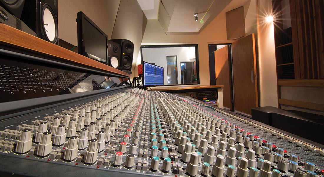 Angle view inside a music recording studio featuring a large analog mixing console with multiple controls, speakers, and sound equipment.