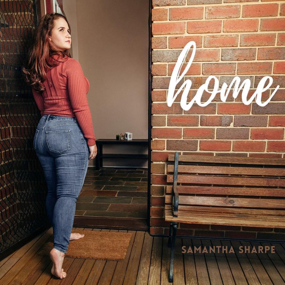 Barefoot woman in jeans and a sweater stands by a brick wall with the word 'home' on it, next to a wooden bench and a door.