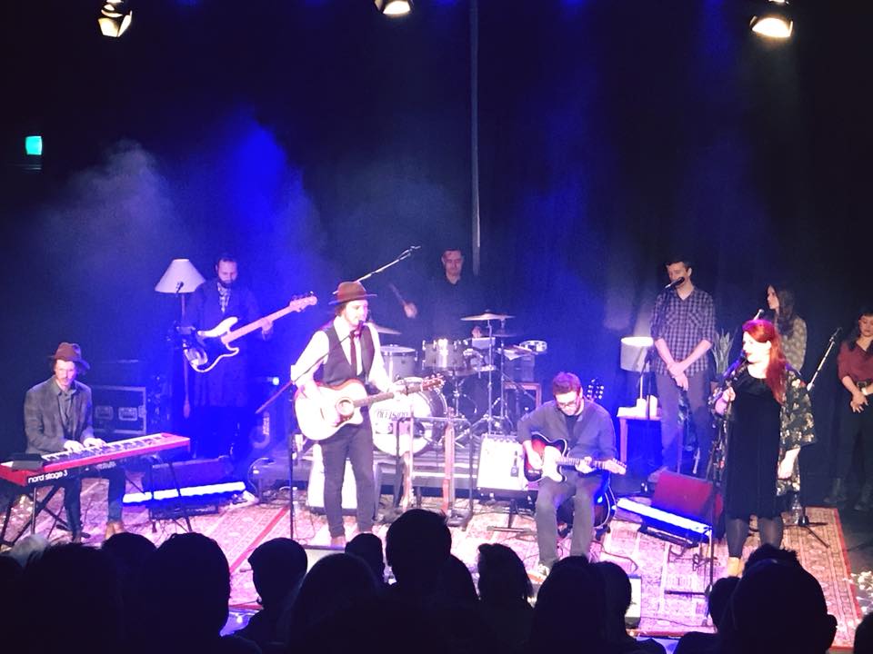 Live performance on stage featuring a band with a singer, musicians playing instruments, and audience members in a dark, indoor setting.