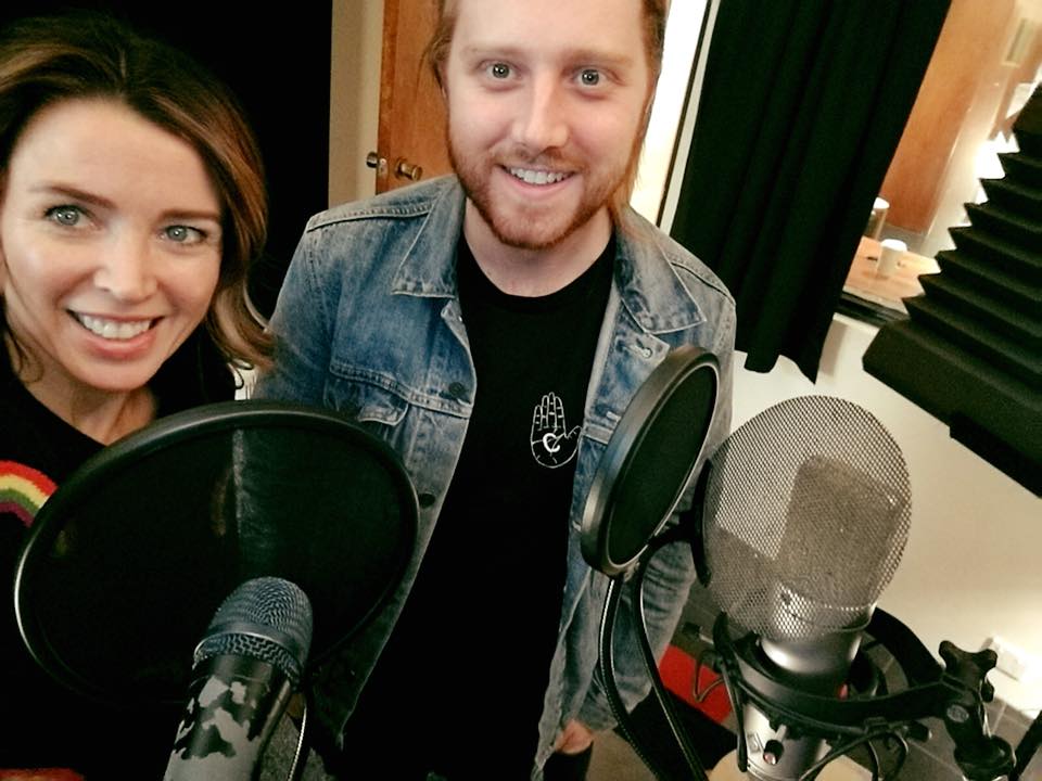 Two adults smiling in a podcast studio with microphones. Woman in black shirt with rainbow, man in denim jacket and black shirt.