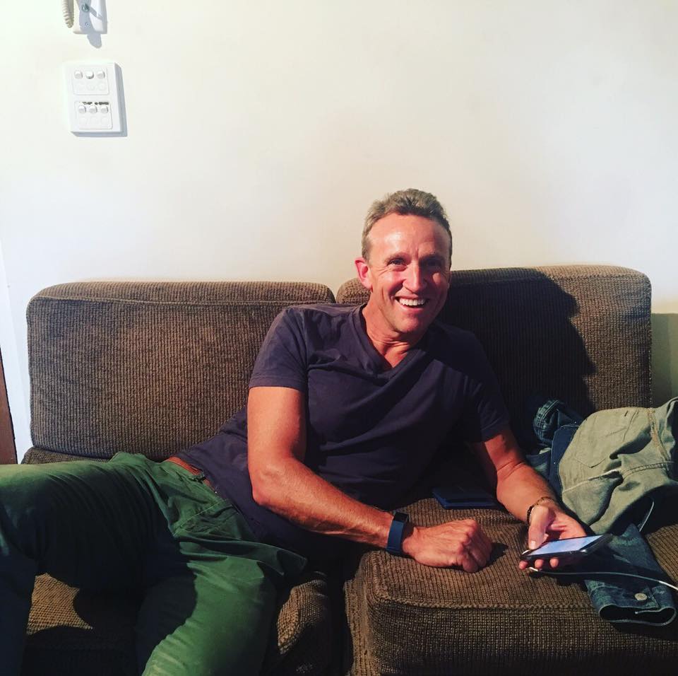 Indoor shot of a smiling adult man relaxing on a brown couch, looking directly at the camera and holding a phone.