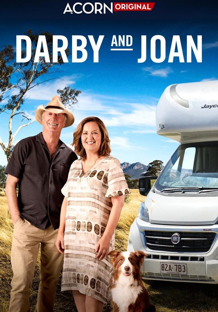 Two people, dog, and RV on a road trip under a sunny sky in an outdoor environment with mountains in the background.