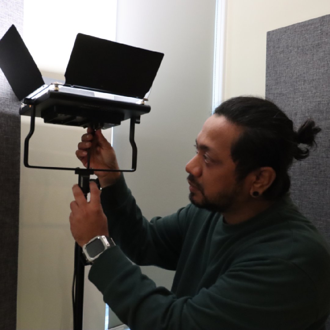 Adult male adjusts a LED panel light on a stand inside a photo studio with acoustic panels and neutral walls, showcasing lighting equipment.