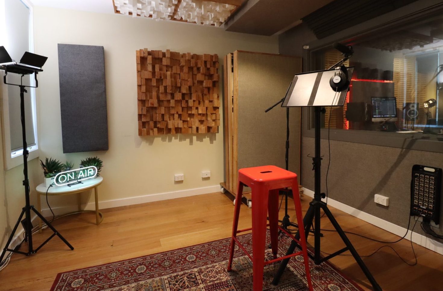 Interior view of a recording studio with acoustic panels, a red stool, microphones on stands, and studio equipment visible through a window.