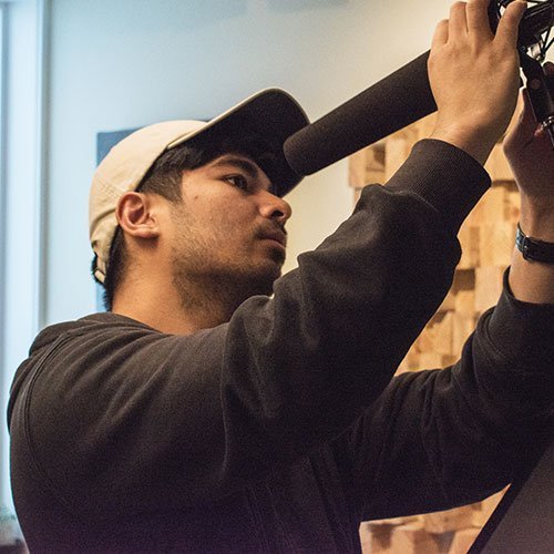 Adult male adjusting microphone in recording studio wearing casual clothing and a baseball cap with soft lighting and wood background.