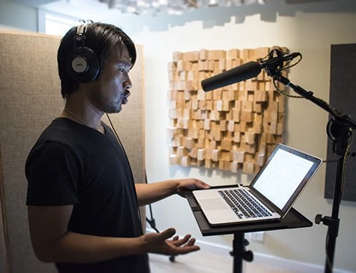 Man recording audio in a sound booth wearing headphones and speaking into a microphone while looking at a laptop screen indoors.
