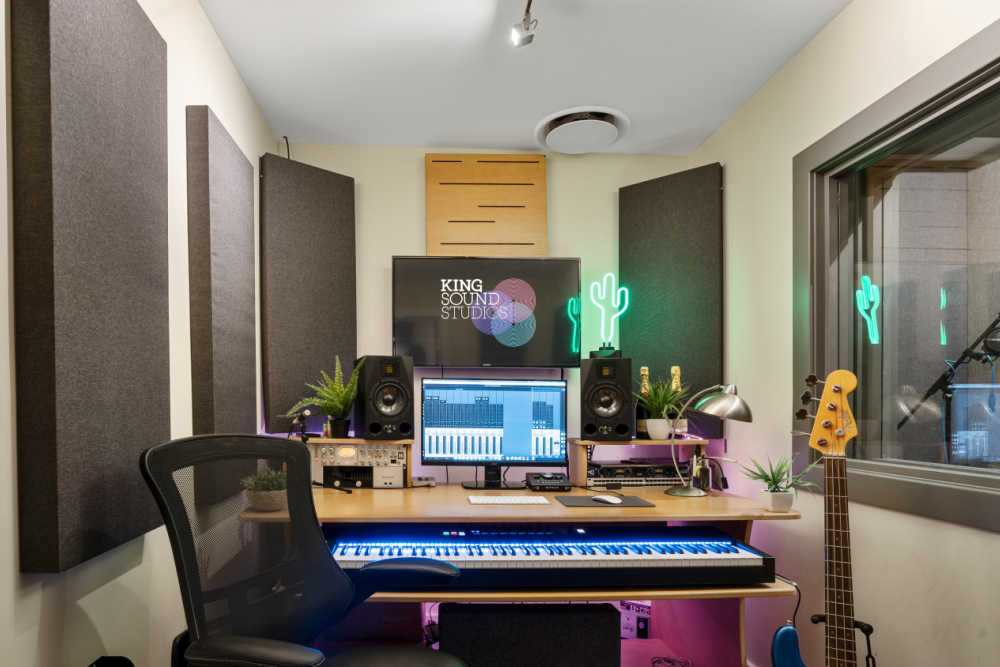 Acoustic music studio with a computer workstation, monitor speakers, keyboard, desk, chair, and a guitar inside a professional recording space.