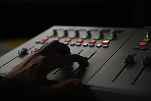 Close-up on a hand operating the sliders of a gray audio mixing console in a dimly lit environment, emphasizing control and sound.