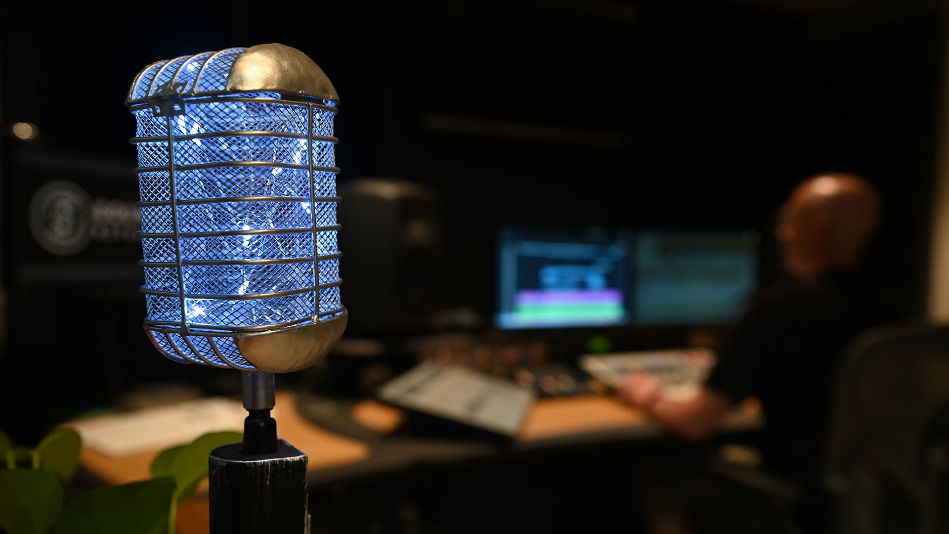 Close-up of decorative microphone with LED lights in a dark interior, with a person working on a computer in the blurred background.