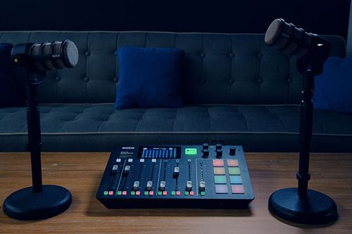 A podcasting setup on a wooden table features two microphones on stands, a RODECaster Pro audio mixer, and a dark grey tufted sofa in the background.