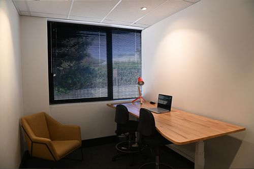 Office space with a wooden desk, laptop, two chairs, and a window with blinds in a modern, minimalist style.