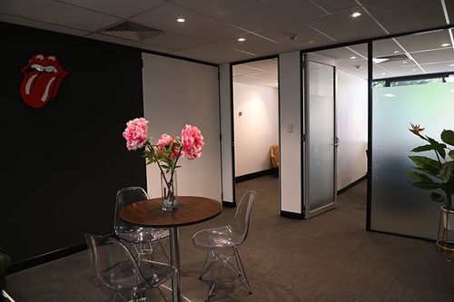 Office lounge area with a brown table topped with pink flowers, surrounded by ghost chairs, and a Rolling Stones wall logo.