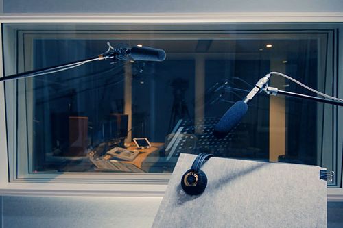 Two microphones and black and gold headphones in a recording studio. The studio has soundproof glass and recording equipment.