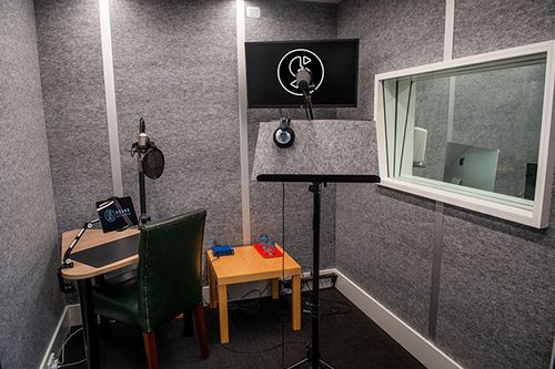 Recording booth with microphone, headphones, music stand, desk, chair, and computer screen visible through glass window, grey color scheme.