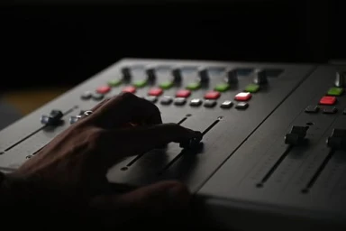 Close-up of a hand adjusting a fader on a professional audio mixing console with illuminated buttons in a dim studio.