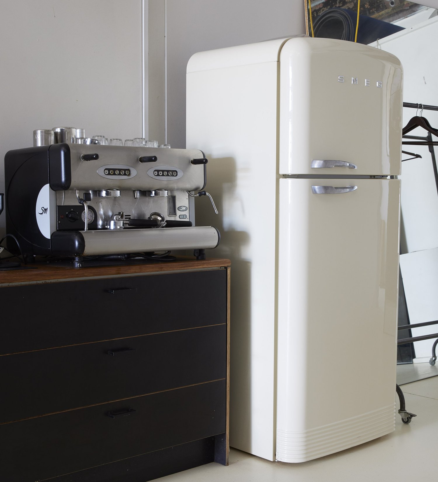 Cream-colored SMEG refrigerator stands near a black dresser with an espresso machine on top in a brightly lit room.