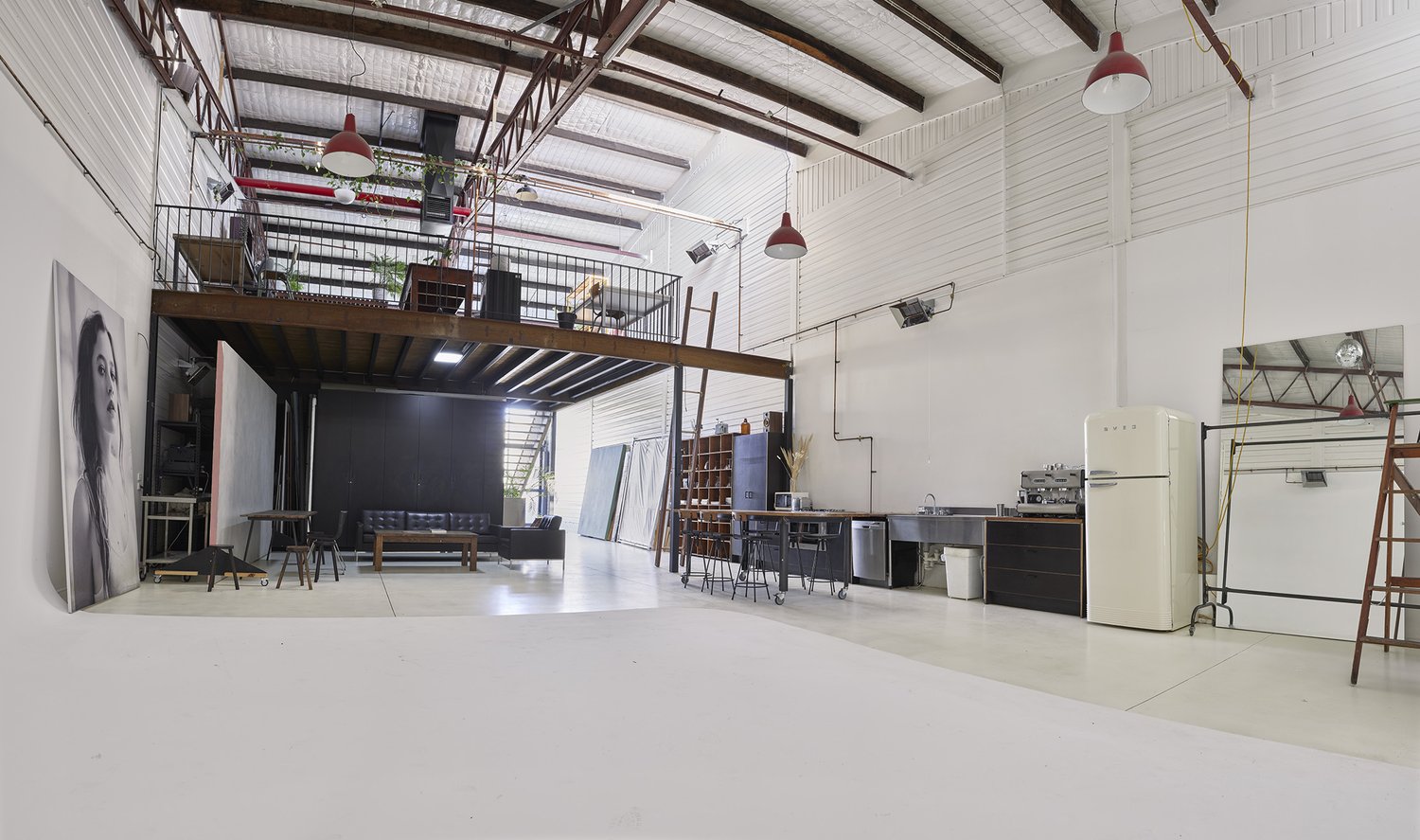 Wide interior shot of a large, bright photo studio featuring a mezzanine level, studio equipment, white walls, and a white floor.